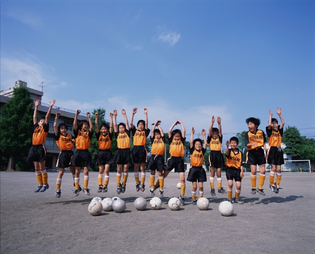 イラスト＆写真のストックフォトwaha（ワーハ）　人物、日本人、元気、健康、子供、スポーツ、小学生、学生、屋外、正面、全身、グループ、仲間、集団、集合、友達、チームメイト、ジャンプ、跳ぶ、バンザイ、手を上げる、サッカーボール、青空、校庭、グラウンド、ユニフォーム、運動、子ども、こども　p5-0120c