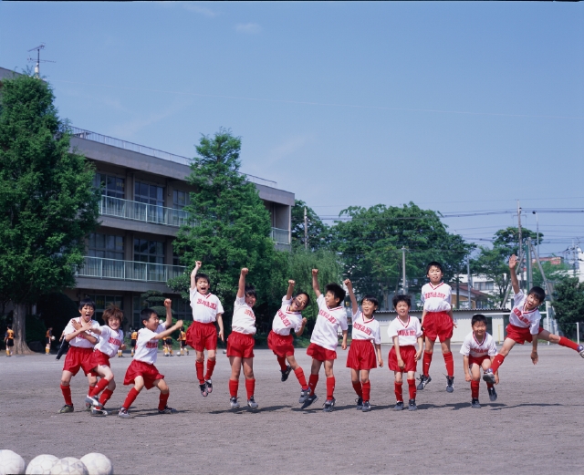 人物 日本人 小学生 学生 スポーツ 子供 元気 健康 屋外 正面 全身 グループ 仲間 集団 集合 友達 チームメイト ジャンプ 跳ぶ ガッツポーズ 拳を上げる 手を上げる サッカーボール 青空 校庭 グラウンド ユニフォーム 叫ぶ 楽しい ふざける