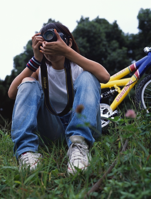 イラスト＆写真のストックフォトwaha（ワーハ）　人物、日本人、若者、自転車、屋外、正面、上半身、ローアングル、女の子、女性、カメラ、写真、覗く、レンズ、好奇心、趣味、リストバンド、草原、原っぱ　p6-0983bv