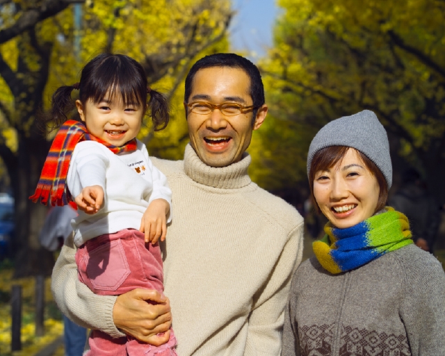 イラスト＆写真のストックフォトwaha（ワーハ）　人物、日本人、公園、秋、家族、屋外、正面、上半身、ポートレート、3人、父、母、子供、娘、愛情、親子、女の子、幼児、笑う、散歩、マフラー、メガネ、抱く、指差し、笑う、笑い、スマイル、季節、四季、子ども、こども　p8-0080bv-data