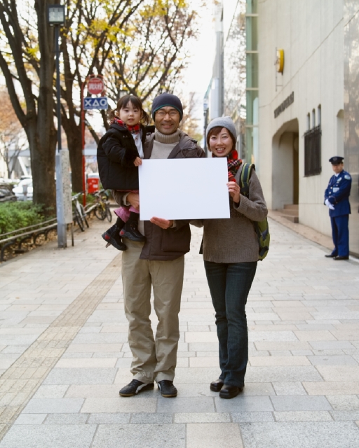 イラスト＆写真のストックフォトwaha（ワーハ）　人物、日本人、メッセージボード、家族、屋外、正面、全身、ポートレート、3人、父、母、子供、娘、愛情、親子、女の子、幼児、笑う、マフラー、メガネ、抱く、街角、ストリート、笑う、笑い、スマイル、街並み、街並、子ども、こども　p8-0143b-data